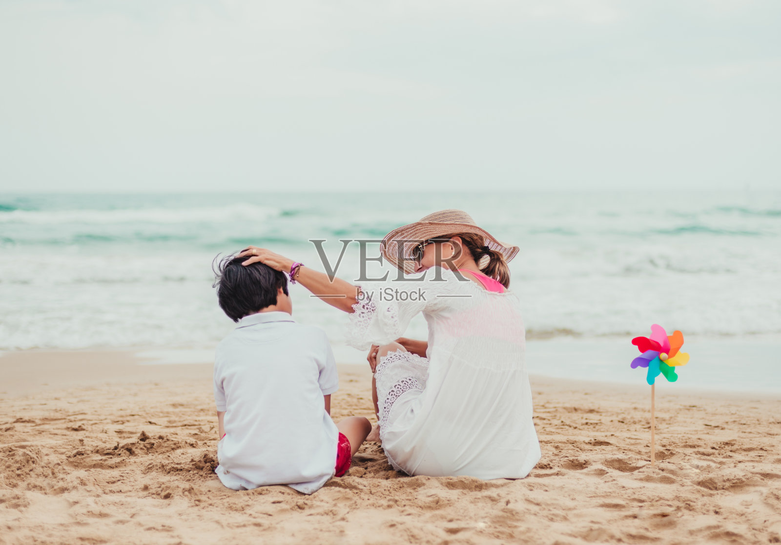 母亲和儿子坐在海边的海滩上。母亲爱抚着她的儿子。他们旁边有一个彩色的风车。单亲家庭的概念。多样性。采用。照片摄影图片