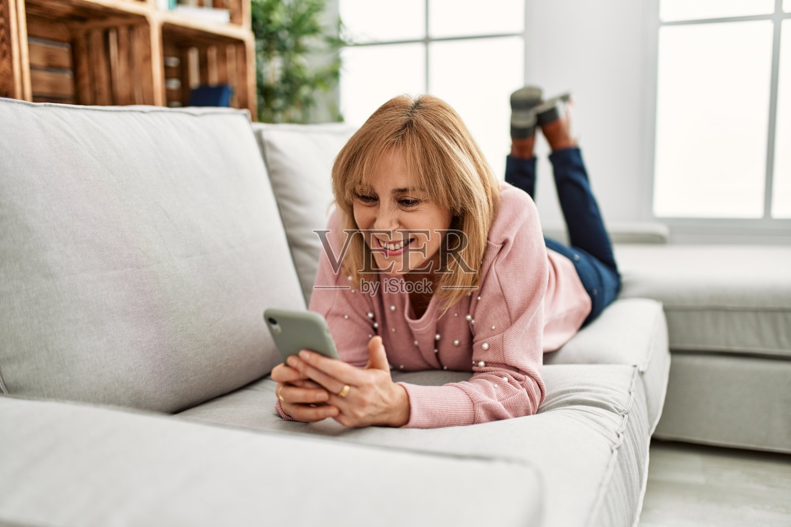 中年金发女人躺在家里的沙发上玩智能手机。照片摄影图片