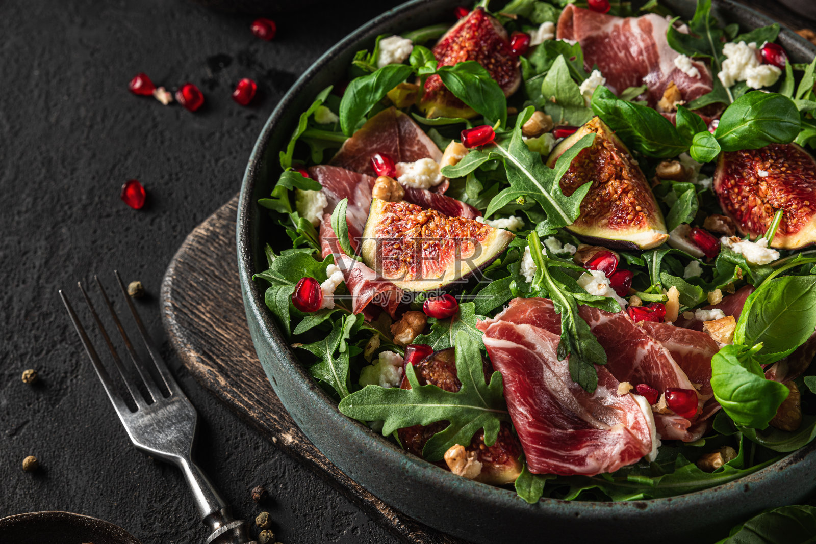 沙拉配无花果，山羊奶酪，意大利熏火腿，水果卷，坚果和石榴，叉子在黑色背景上。近距离照片摄影图片