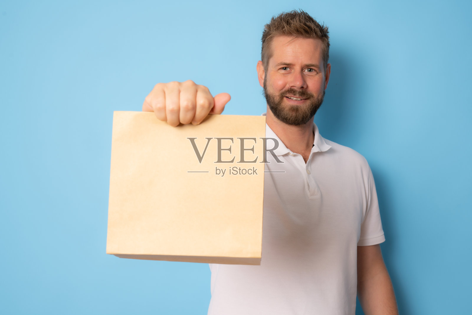 年轻微笑的男子展示棕色清楚空白的手工纸外卖袋模拟孤立在蓝色背景。照片摄影图片