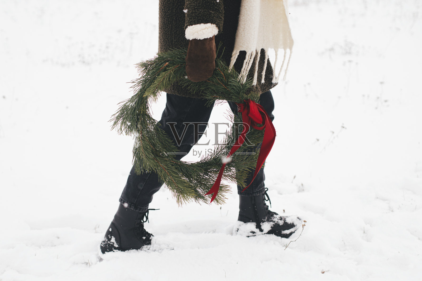 圣诞快乐!冬季公园里戴着圣诞花环的时髦女人。年轻女性在舒适的手套和外套与圣诞花环红色蝴蝶结在雪域冬季，裁剪的观点。照片摄影图片