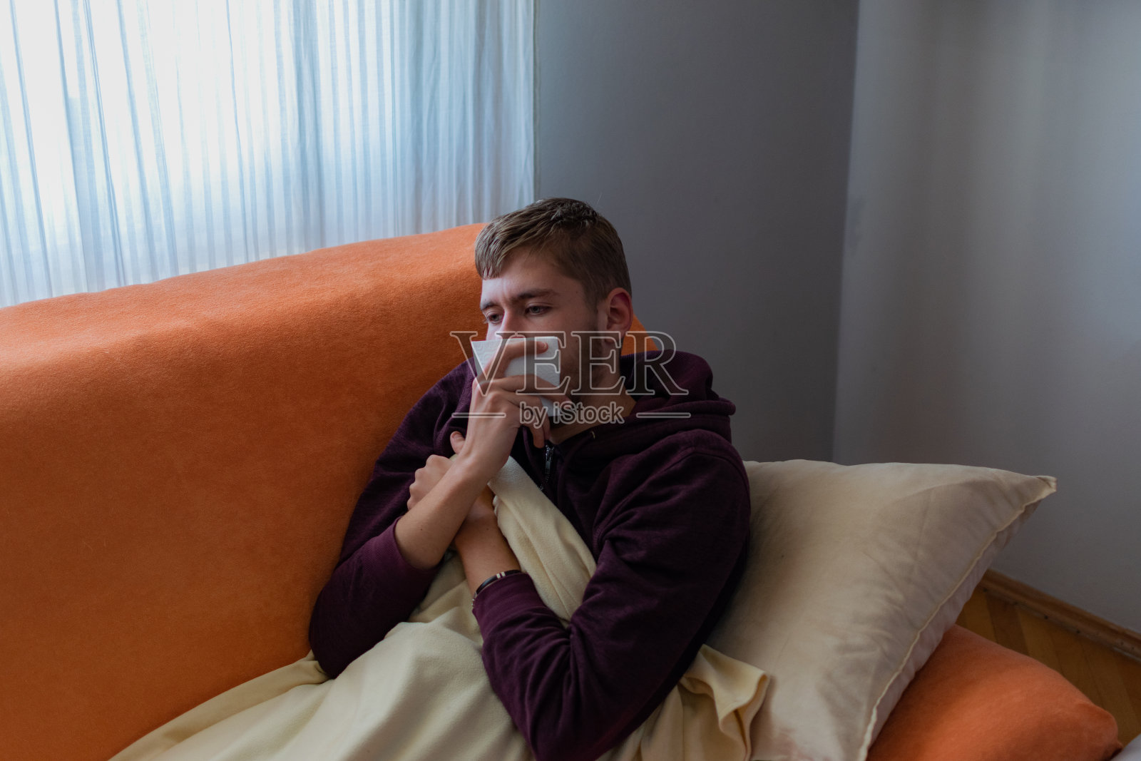 一个生病的年轻人坐在家里，喝着茶，在客厅里休息，吃着药和维生素，量着体温，盖着毯子躺在床上看电视。照片摄影图片