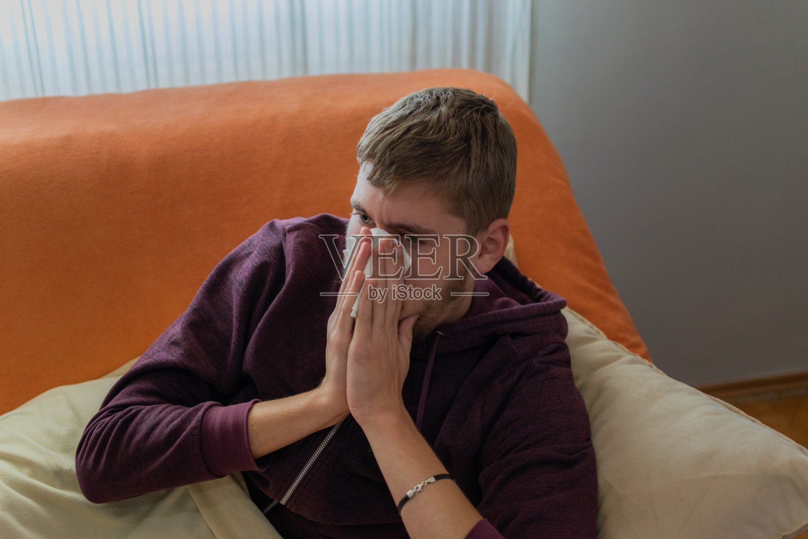 一个生病的年轻人坐在家里，喝着茶，在客厅里休息，吃着药和维生素，量着体温，盖着毯子躺在床上看电视。照片摄影图片