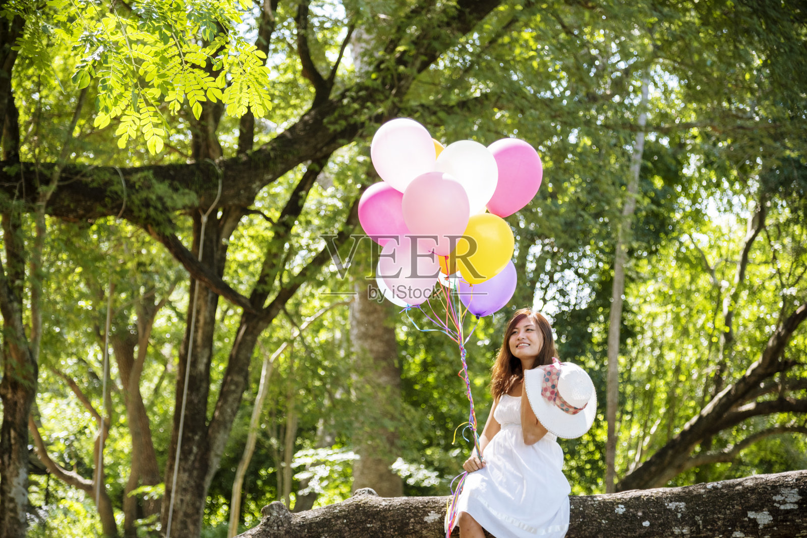 欢快的美女拿着气球坐在绿色公园的大树下，心情舒畅。女人手握充满活力的气球在生日派对上玩快乐的时间夏天在户外的阳光照片摄影图片