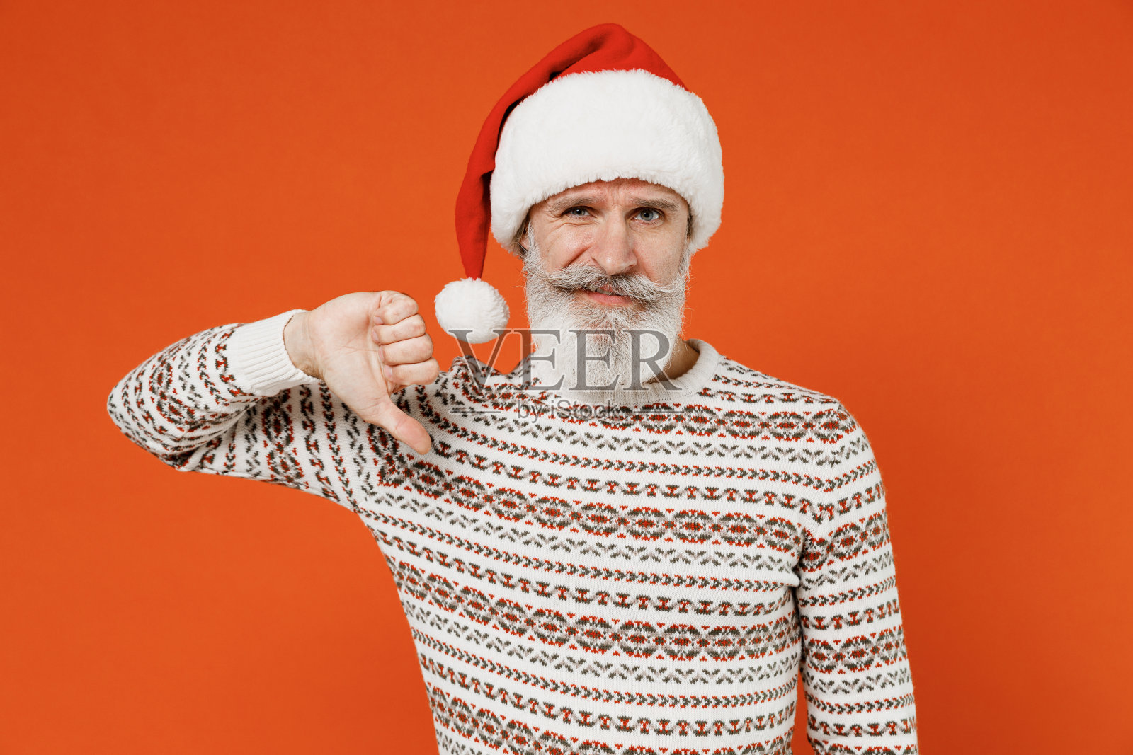 年迈沮丧，满脸胡须，情绪不稳，愤愤不平，不安的圣诞老人，50多岁，戴着圣诞帽，穿着毛衣，摆着姿势看着摄像机，拇指朝下，不喜欢的手势孤立在普通的橙色背景摄影棚照片摄影图片