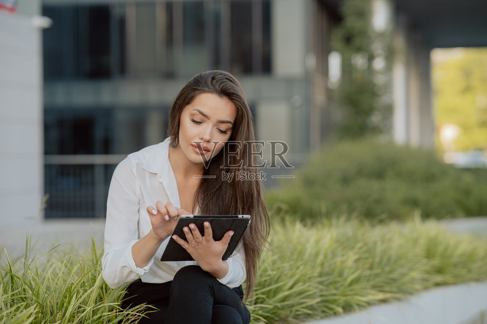 一个微笑的女人，公司员工，秘书，坐在公司大楼前休息，一堵植物墙，一个黑发的女人手里拿着平板电脑，浏览社交媒体，浏览互联网照片摄影图片