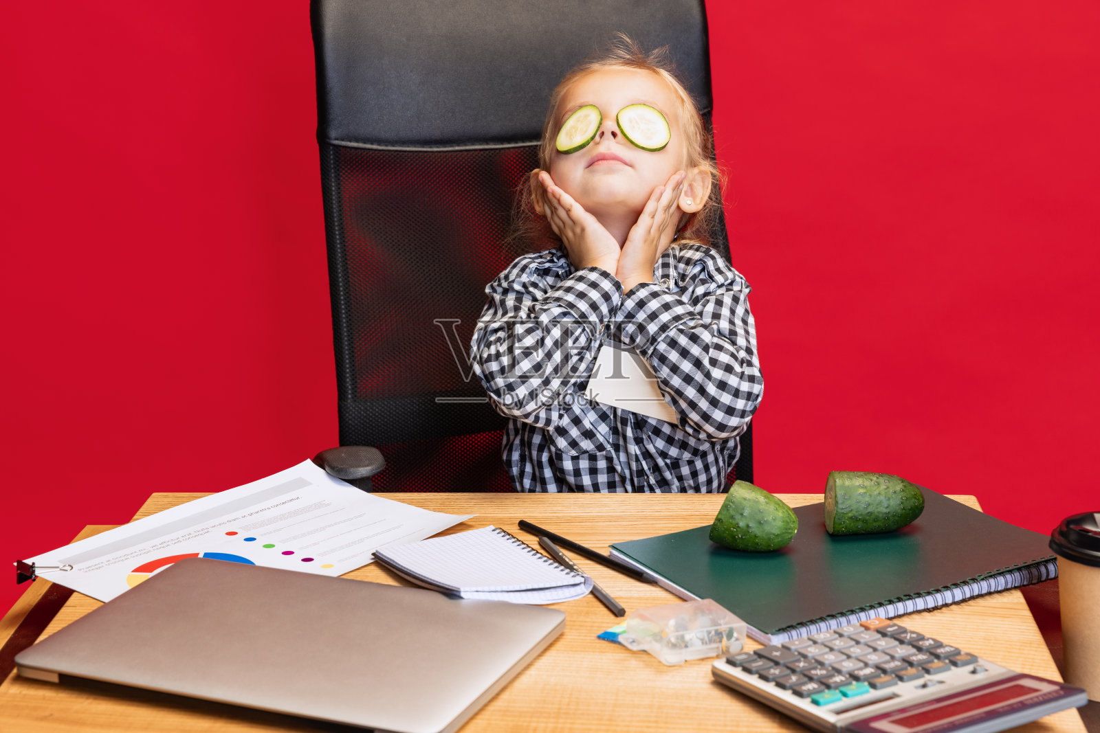 可爱的小女孩坐在桌子旁的裁剪肖像，用红色背景上孤立的黄瓜制作眼罩。照顾照片摄影图片