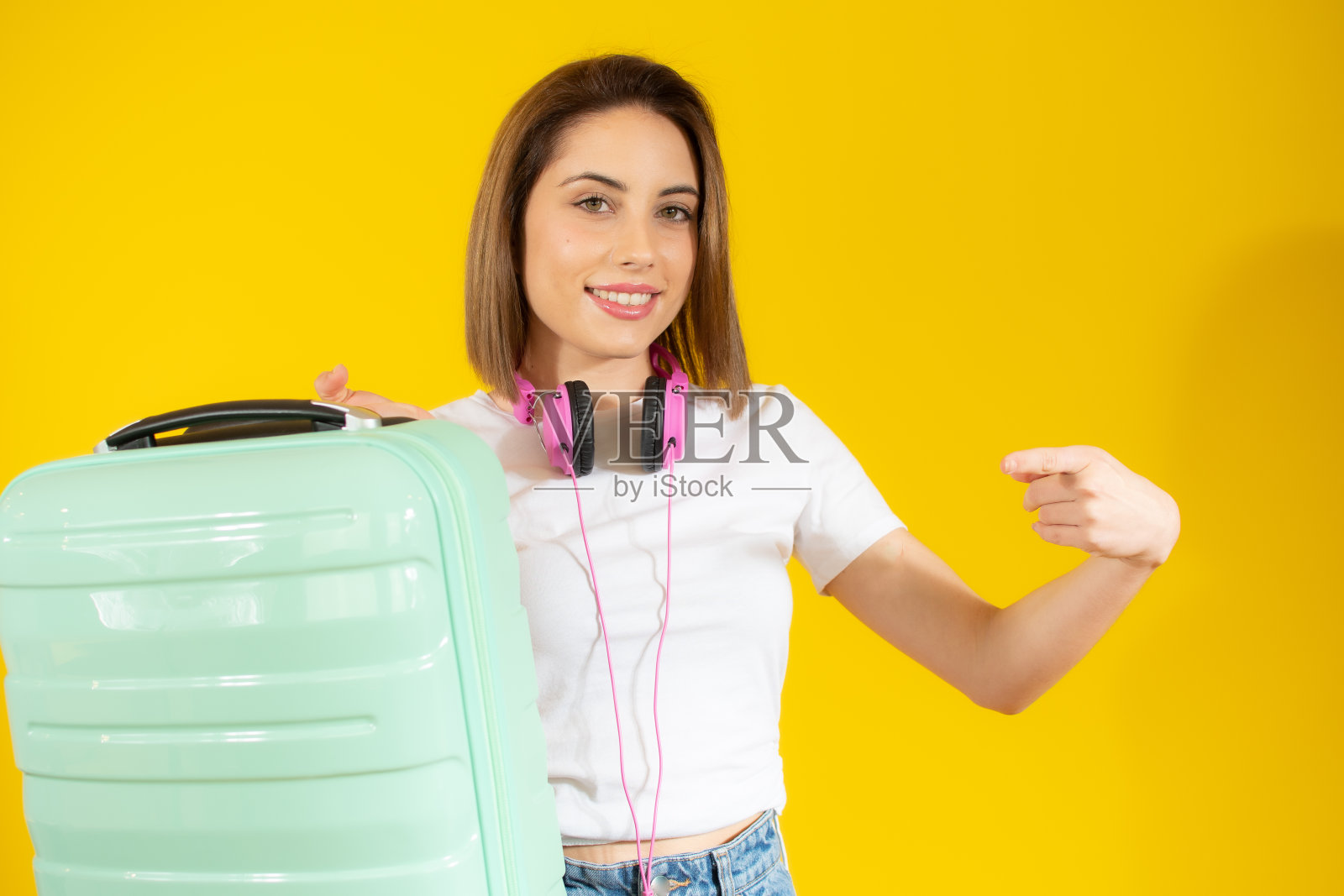 年轻快乐的美丽女人戴着耳机，黄色背景上拿着黄色手提箱照片摄影图片