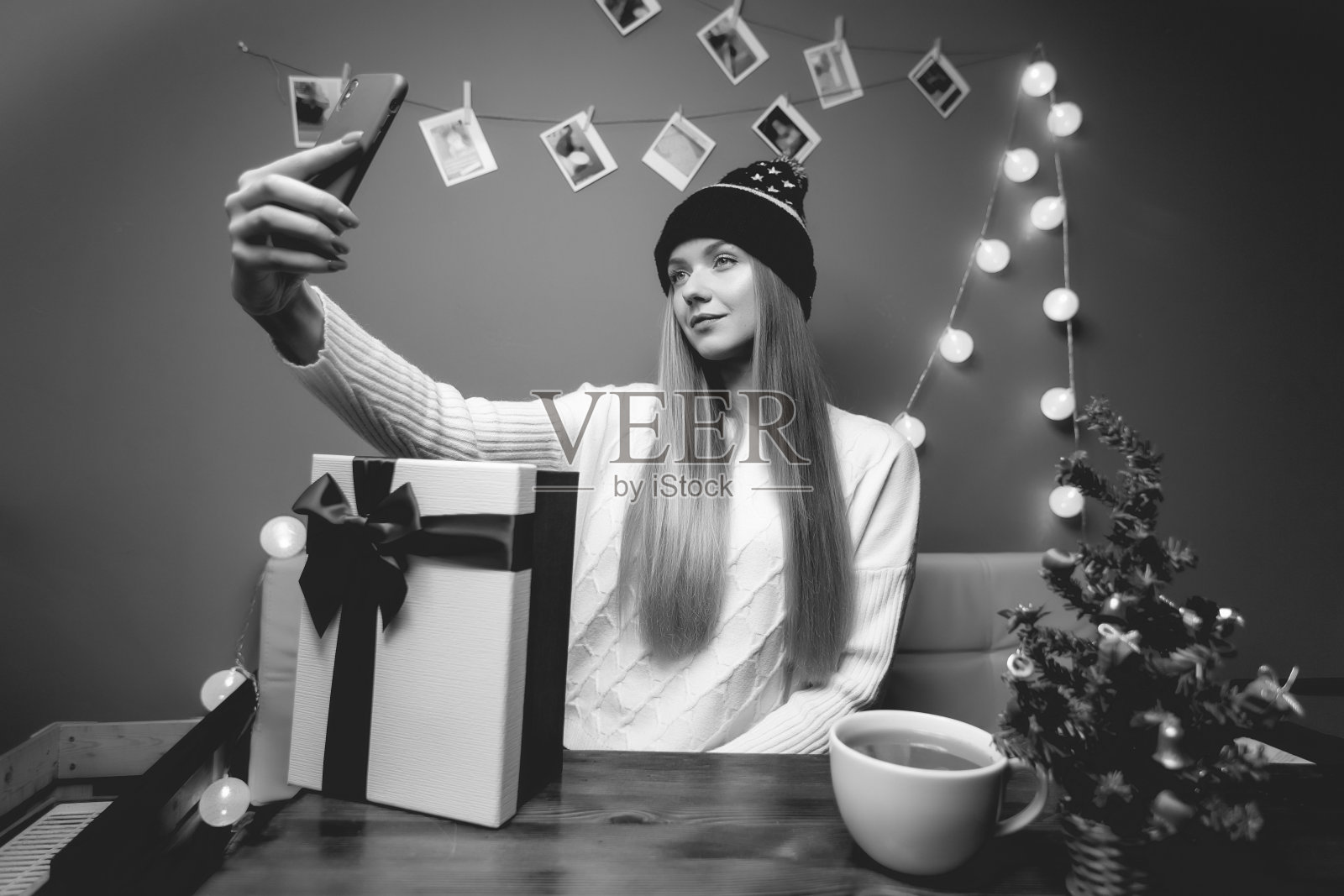 室内女性圣诞肖像。一个女人用手机打视频电话和自拍。冠状病毒和社交距离概念。新年黑白照片与圣诞树和礼盒照片摄影图片