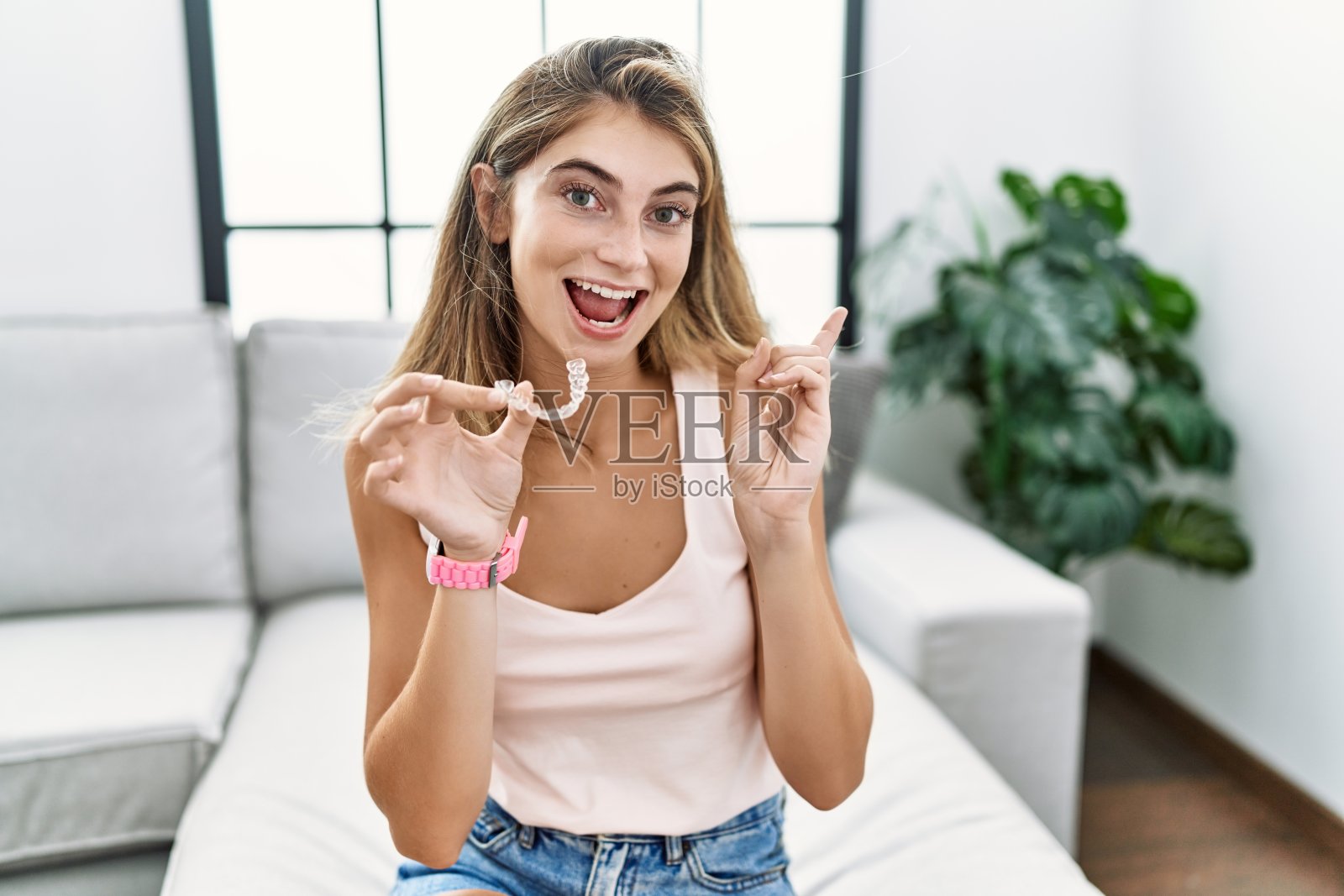 年轻的金发女人拿着隐形矫正器，脸上带着大大的微笑，用手指指着一边看着摄像机。照片摄影图片