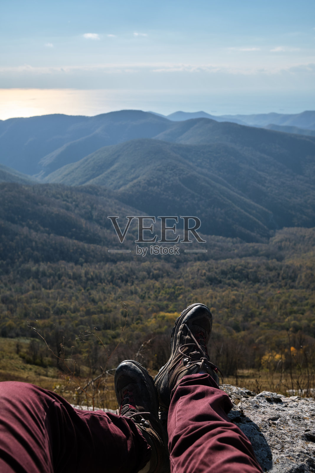第一个人的观点。旅行和探索世界。人类旅行者坐在悬崖上，穿着靴子向前伸展双脚，欣赏秋天的森林和山峰。照片摄影图片