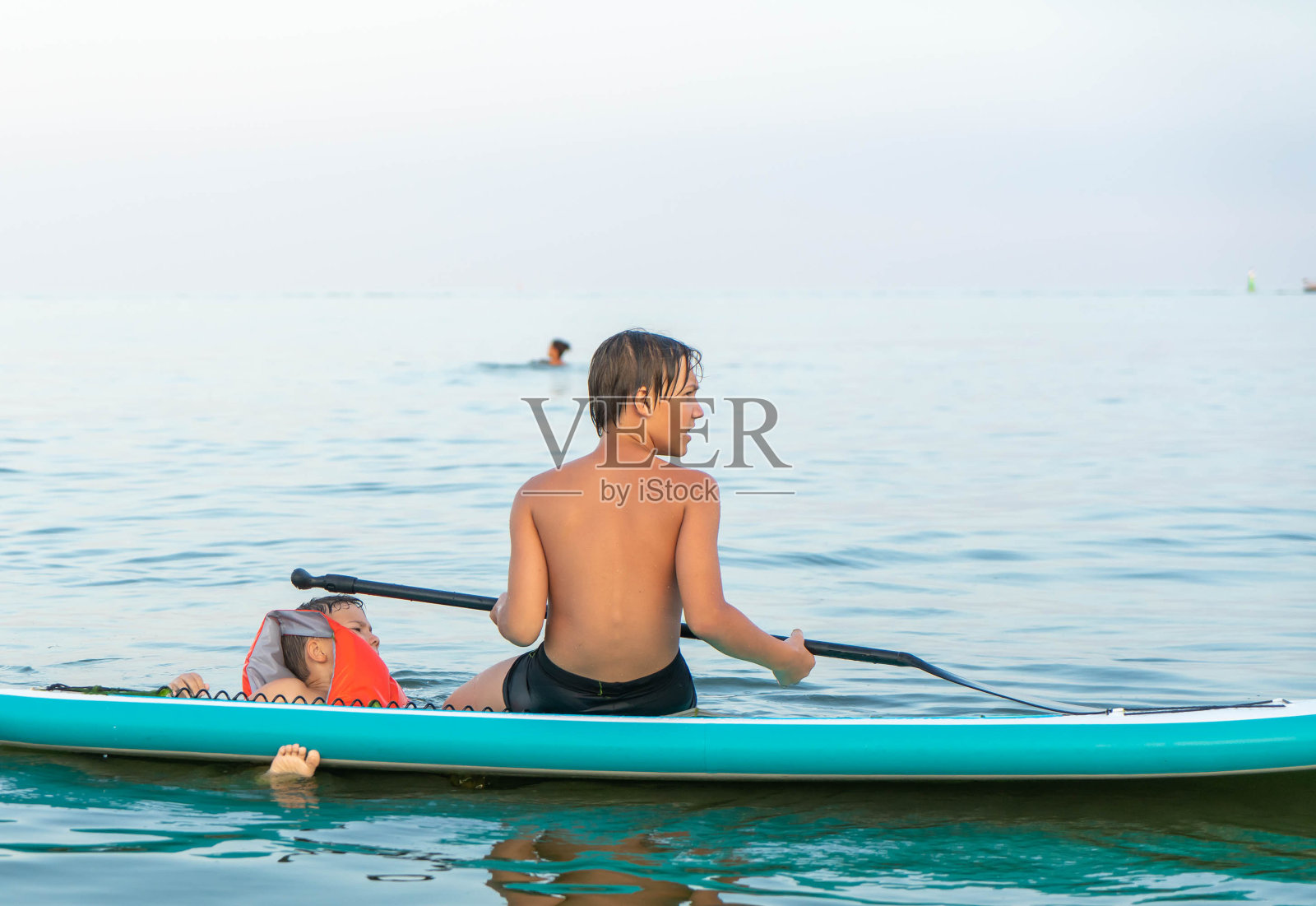两兄弟在海边游泳。一个12岁的男孩在吃晚餐板和一个6岁的孩子穿着救生衣。照片摄影图片