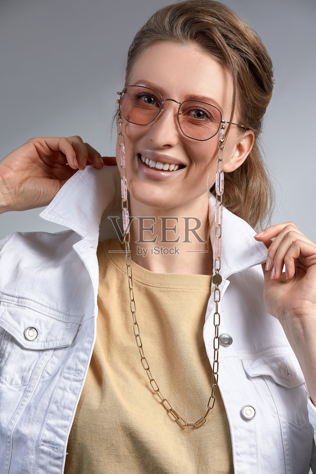 一个美丽的穿着时髦的中年妇女，戴着时髦的眼镜，微笑着对着镜头摆姿势，一个35-45岁的金发女人，灰色背景下戴着眼镜，皮肤健康照片摄影图片