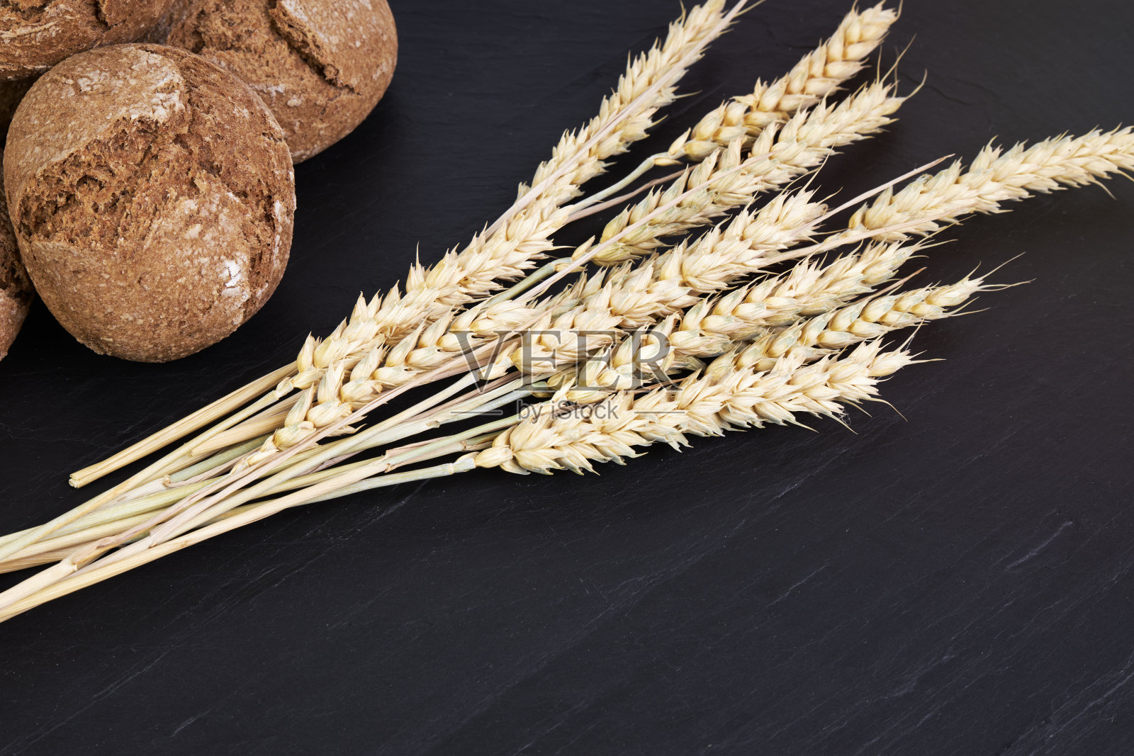 黑麦面包卷旁边的麦穗在一块黑色的石板上。健康食品的概念照片摄影图片