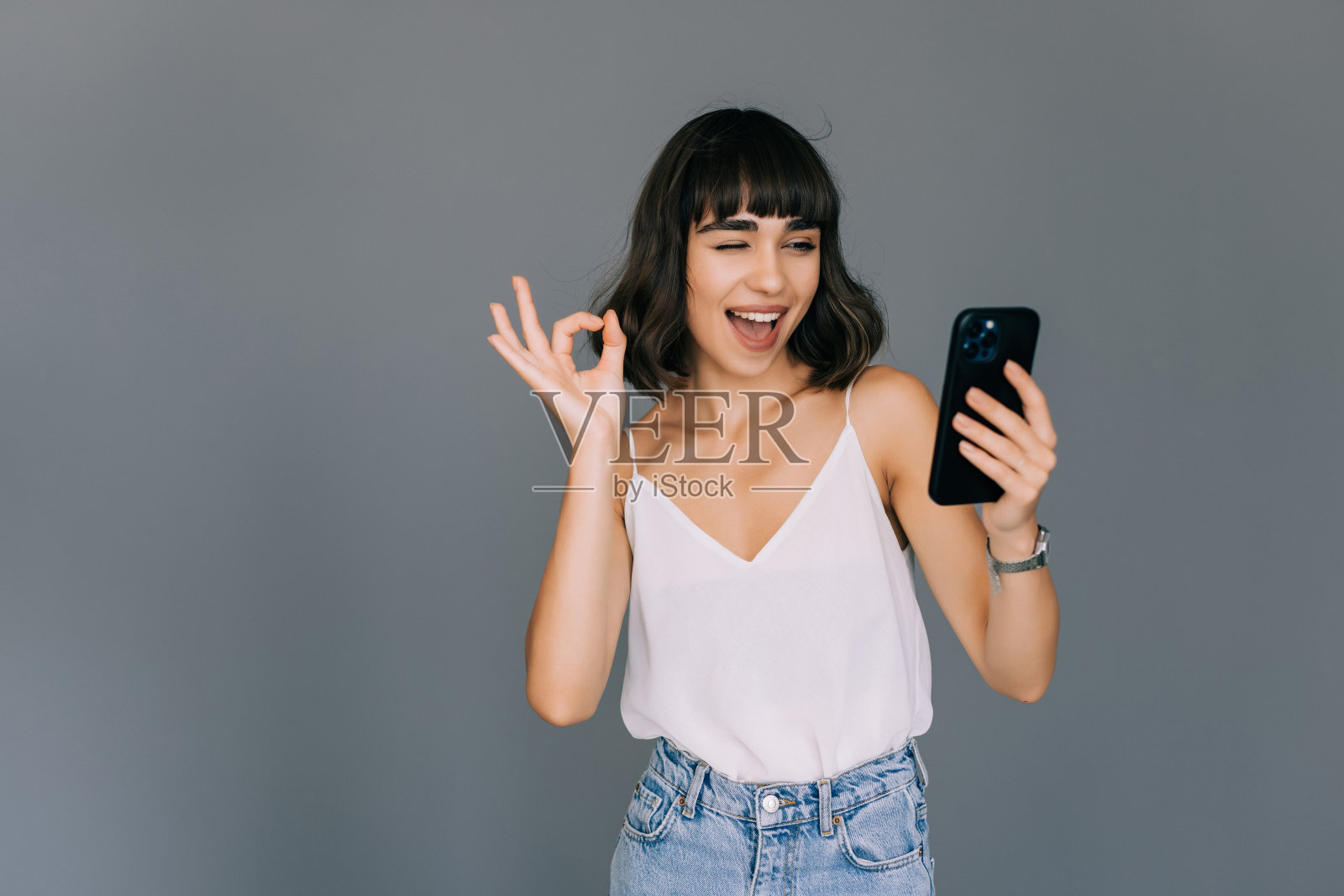 有魅力的年轻女子在灰色背景下拿着手机，做出ok的手势照片摄影图片