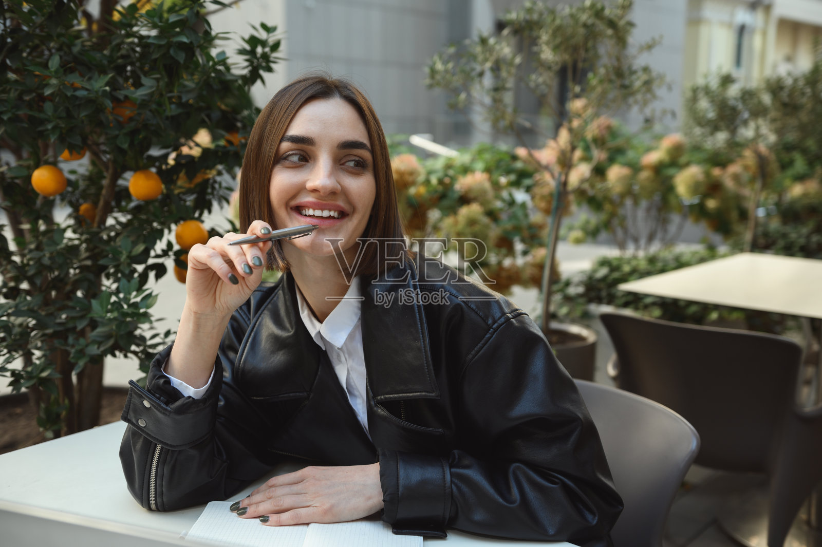 时尚迷人的黑发美女的生活方式肖像在生态皮夹克握笔靠近嘴唇，微笑看着一边坐在咖啡馆的阳台上，背景是橘子树照片摄影图片
