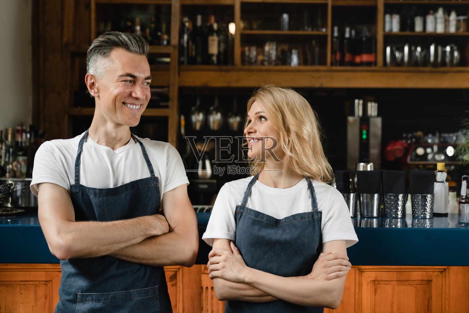 自信成熟的白人小企业主，服务员，穿着蓝色围裙的调酒师，在餐厅的吧台边交叉双臂微笑着看着对方照片摄影图片
