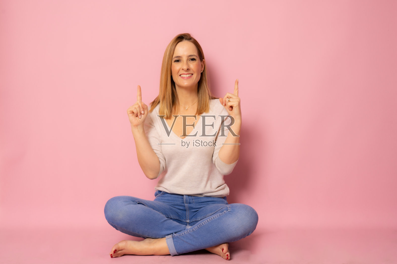 快乐的年轻女子坐在地板上手指指向孤立的粉红色背景。照片摄影图片