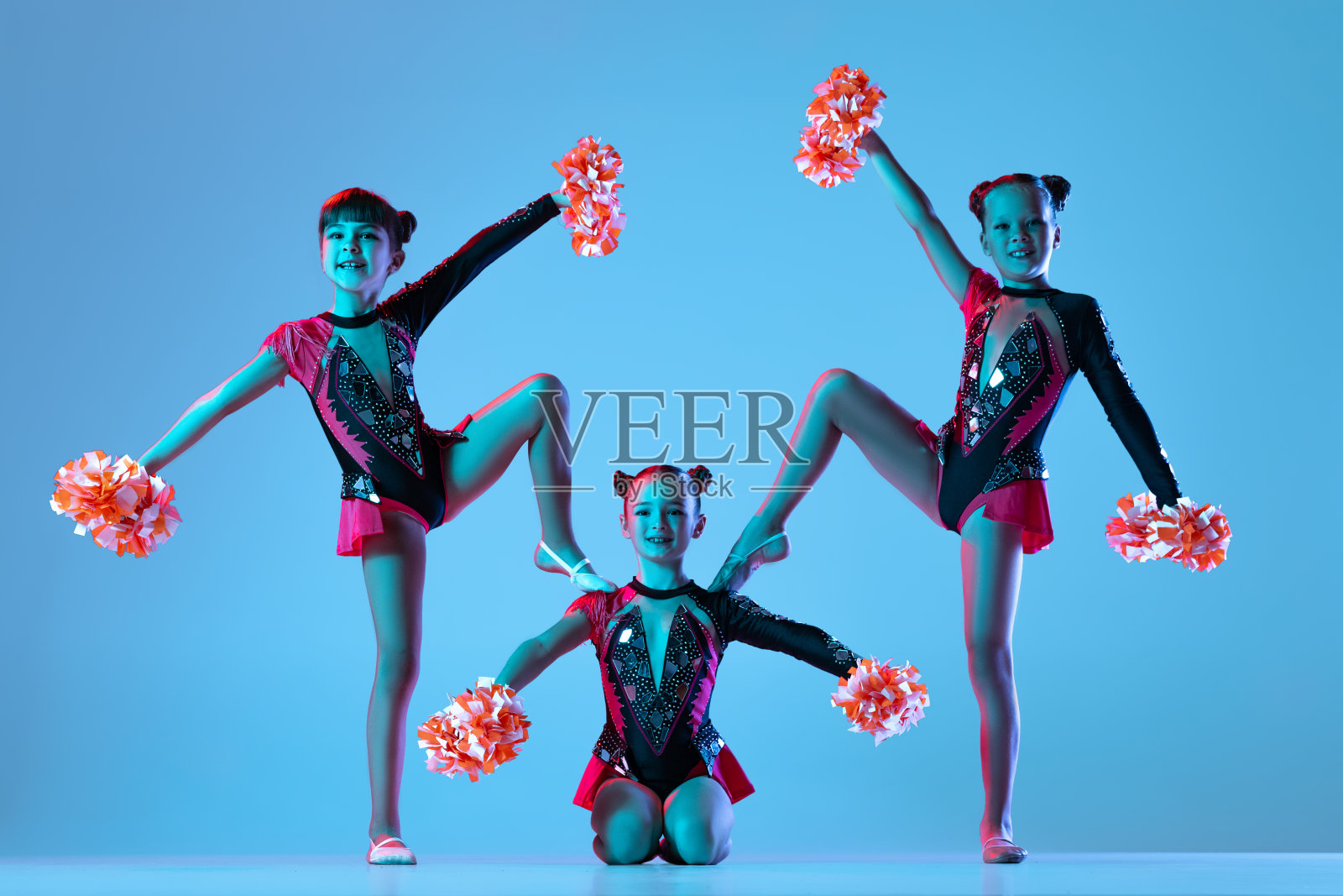三个女孩的全身肖像，啦啦队长训练穿着五颜六色的服装与绒球孤立在蓝色背景霓虹灯照片摄影图片