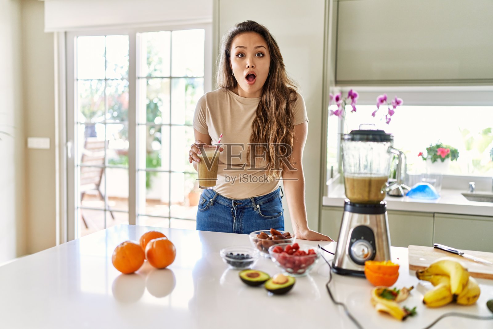 一个年轻漂亮的黑发女人在厨房里喝着一杯冰沙，她张着嘴惊讶地看着，脸上露出难以置信的表情照片摄影图片