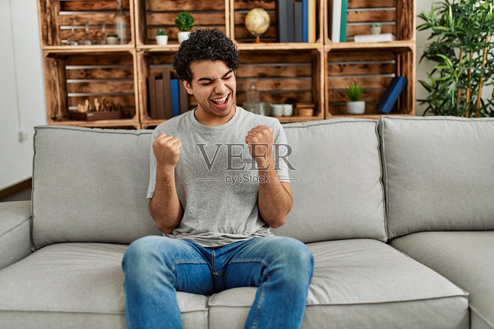 年轻的西班牙裔男子穿着便服坐在家里的沙发上，非常高兴和兴奋地举起双臂，做着胜利者的手势，微笑着大喊着成功。庆祝活动的概念。照片摄影图片