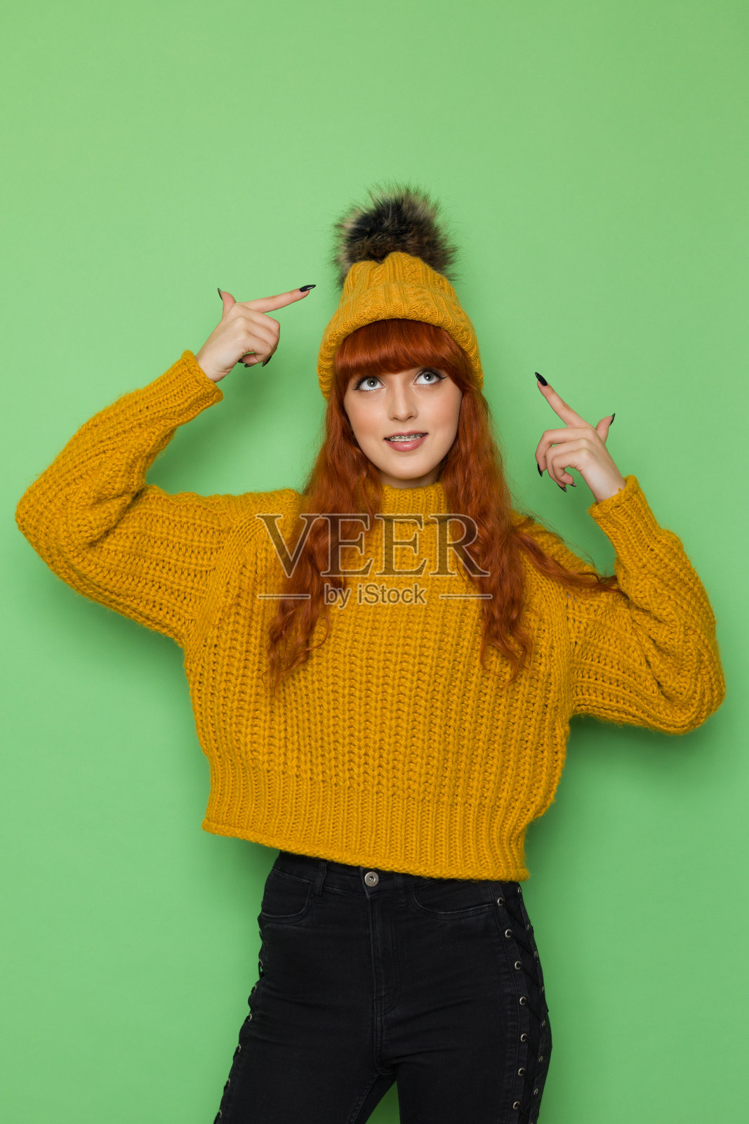 一个可爱的年轻女子正用绒球指着她的帽子，向上看照片摄影图片