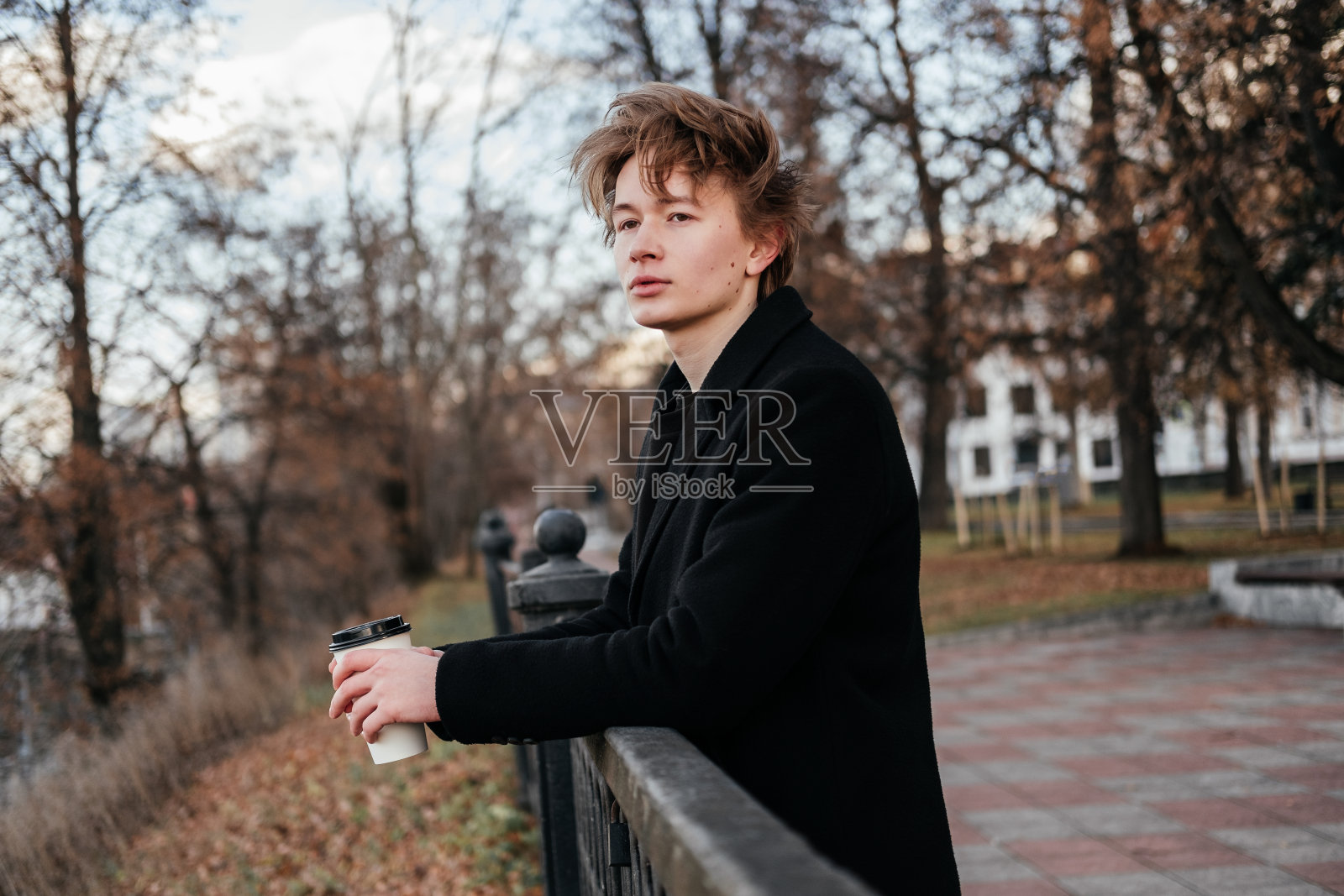 一个身穿黑色外套，手里拿着咖啡的年轻人站在城市的街道上，靠在栅栏上，望着别处。英俊的男人在等人。在秋天的公园里呼吸新鲜空气。照片摄影图片