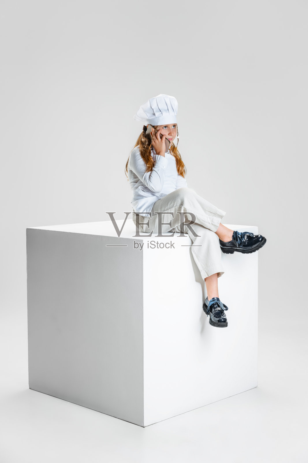 摄影的小女孩，学生穿着白色的厨师制服和巨大的厨师帽坐在巨大的盒子孤立在白色的工作室背景。照片摄影图片