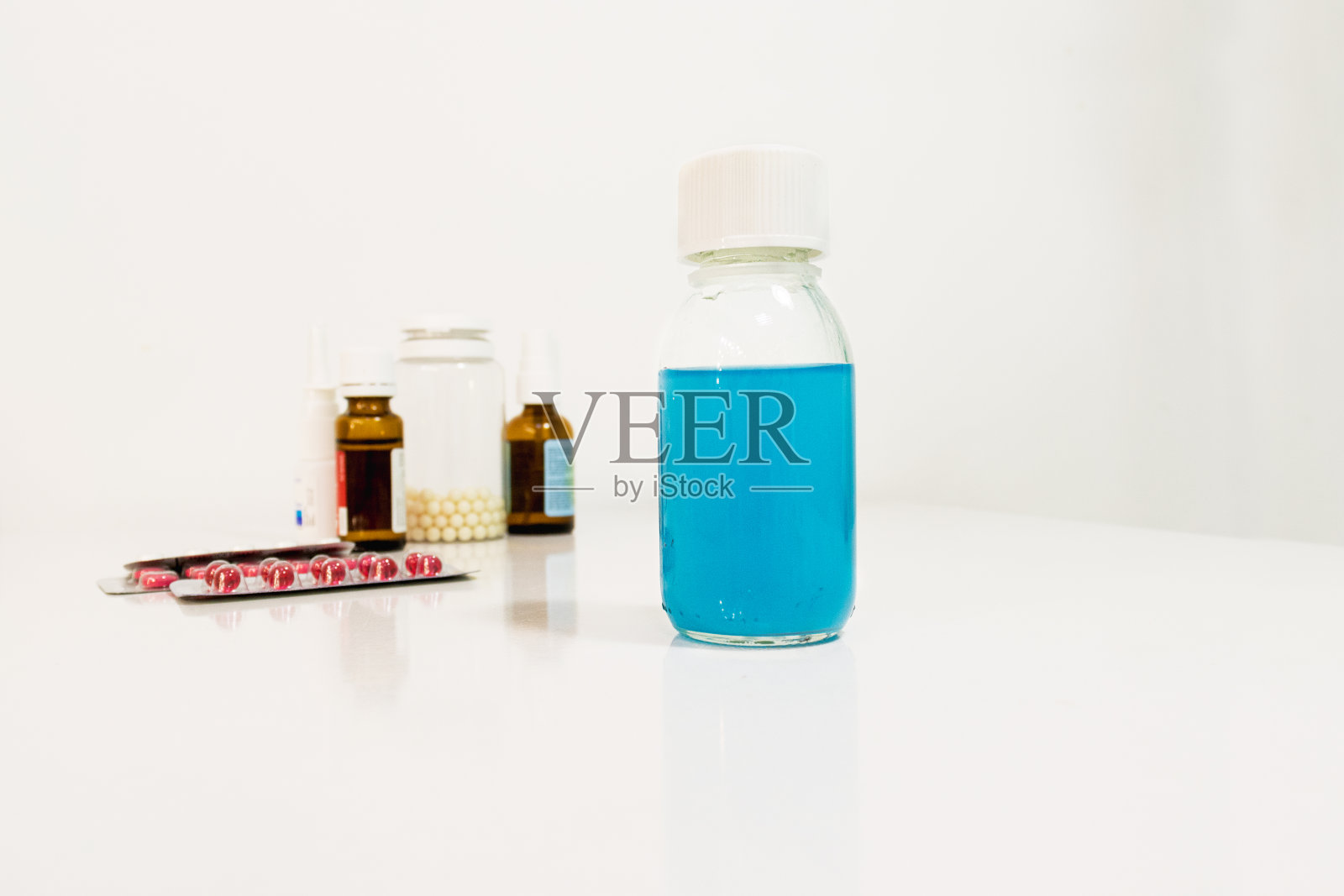 蓝色的医用糖浆放在桌子上，背景是散焦的药丸和液滴。灰色的背景。有选择性的重点。照片摄影图片