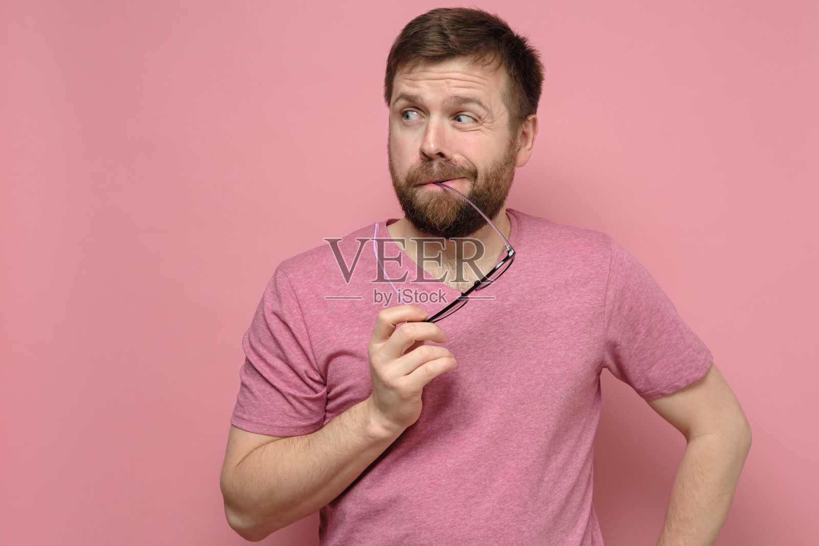 满脸胡须、身穿粉色t恤的男子怀疑有人，他嘴里衔着眼镜，看起来怪怪的。照片摄影图片