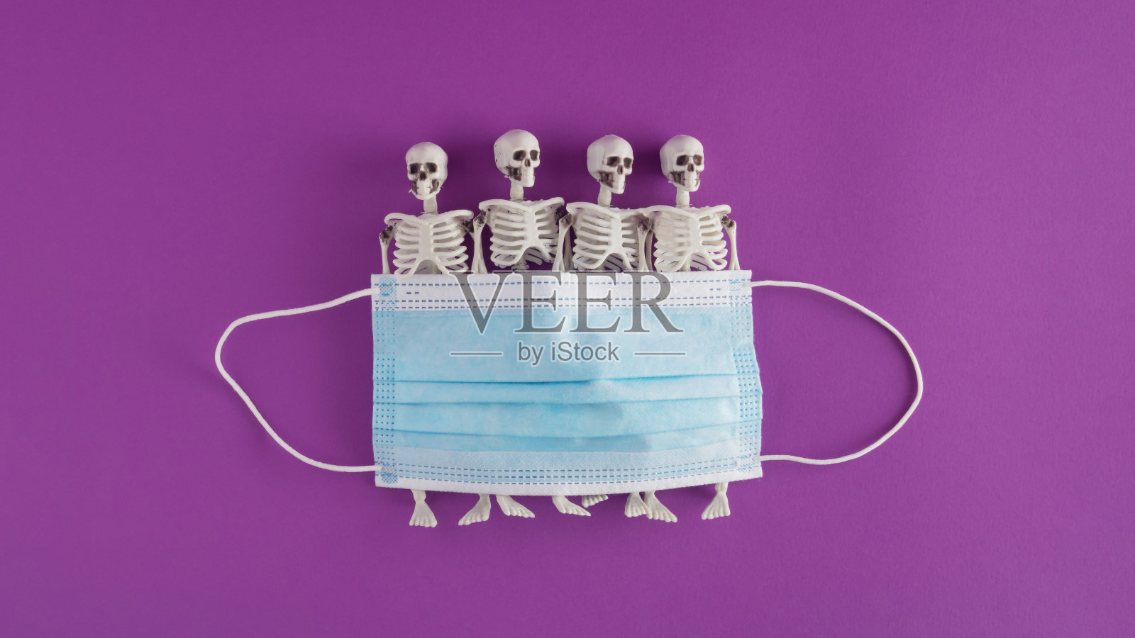 在紫色背景下由面具和骷髅组成的万圣节创意布置。最小的 大流行和新冠概念。万圣节派对的灵感。平的。照片摄影图片