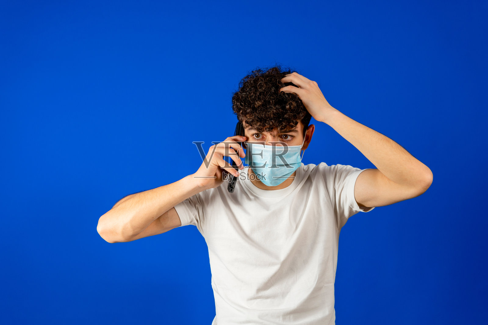 卷发的年轻男子戴着外科口罩以保护自己免受冠状病毒的侵袭，在蓝色录音室背景下愤怒地讲着手机。正常的新概念照片摄影图片
