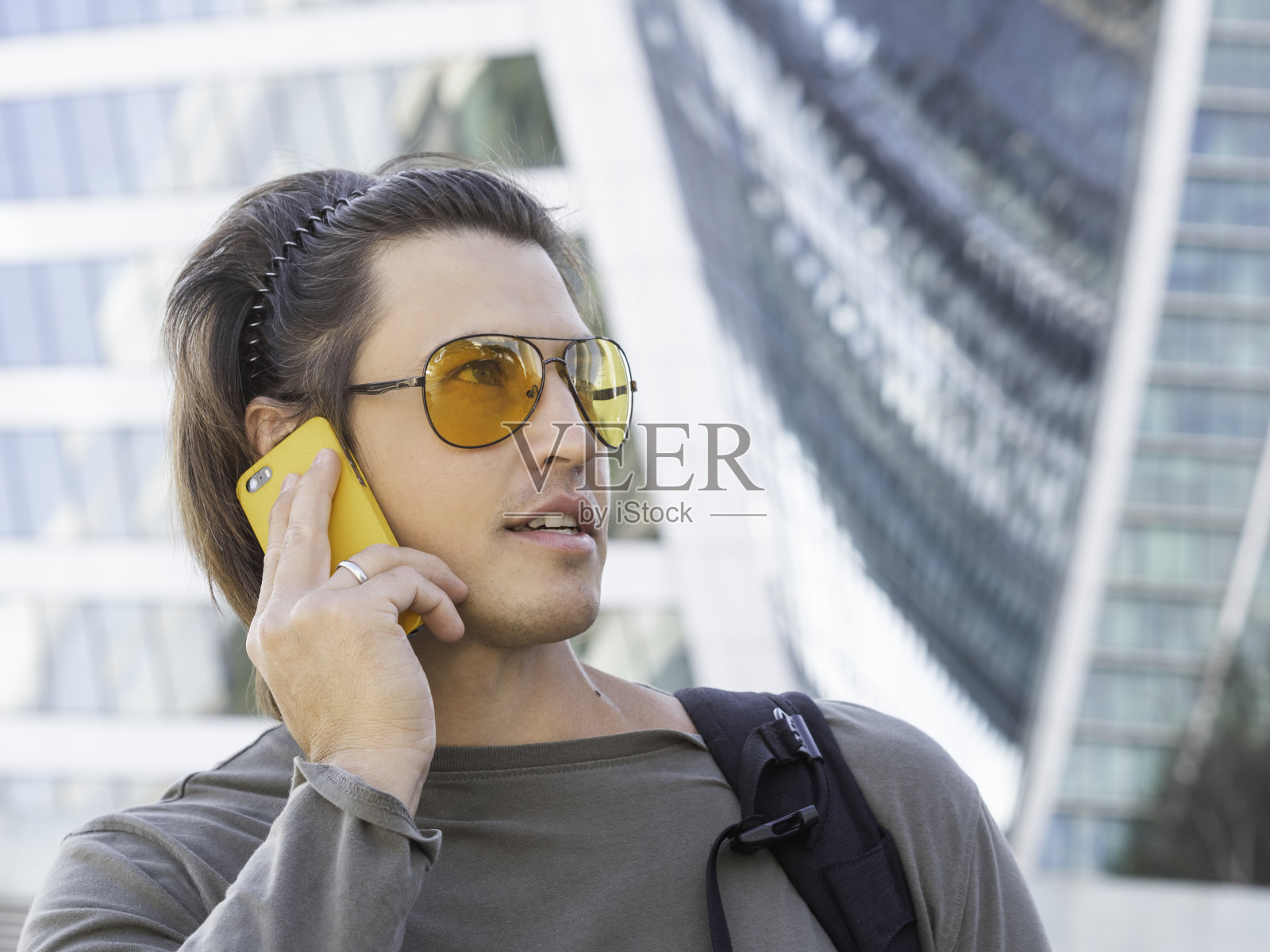 戴着黄色眼镜的年轻人在大城市的背景下用智能手机说话。玻璃幕墙的现代建筑。千禧年的生活方式。照片摄影图片
