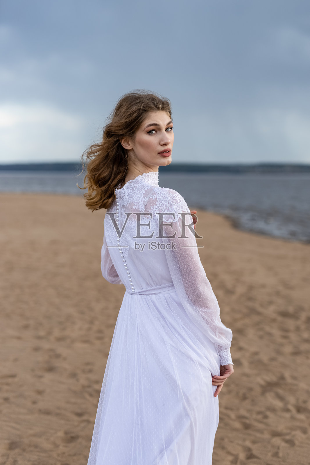 快乐的年轻女孩穿着白色的裙子站在河岸上。河上有暴风雨的天空照片摄影图片