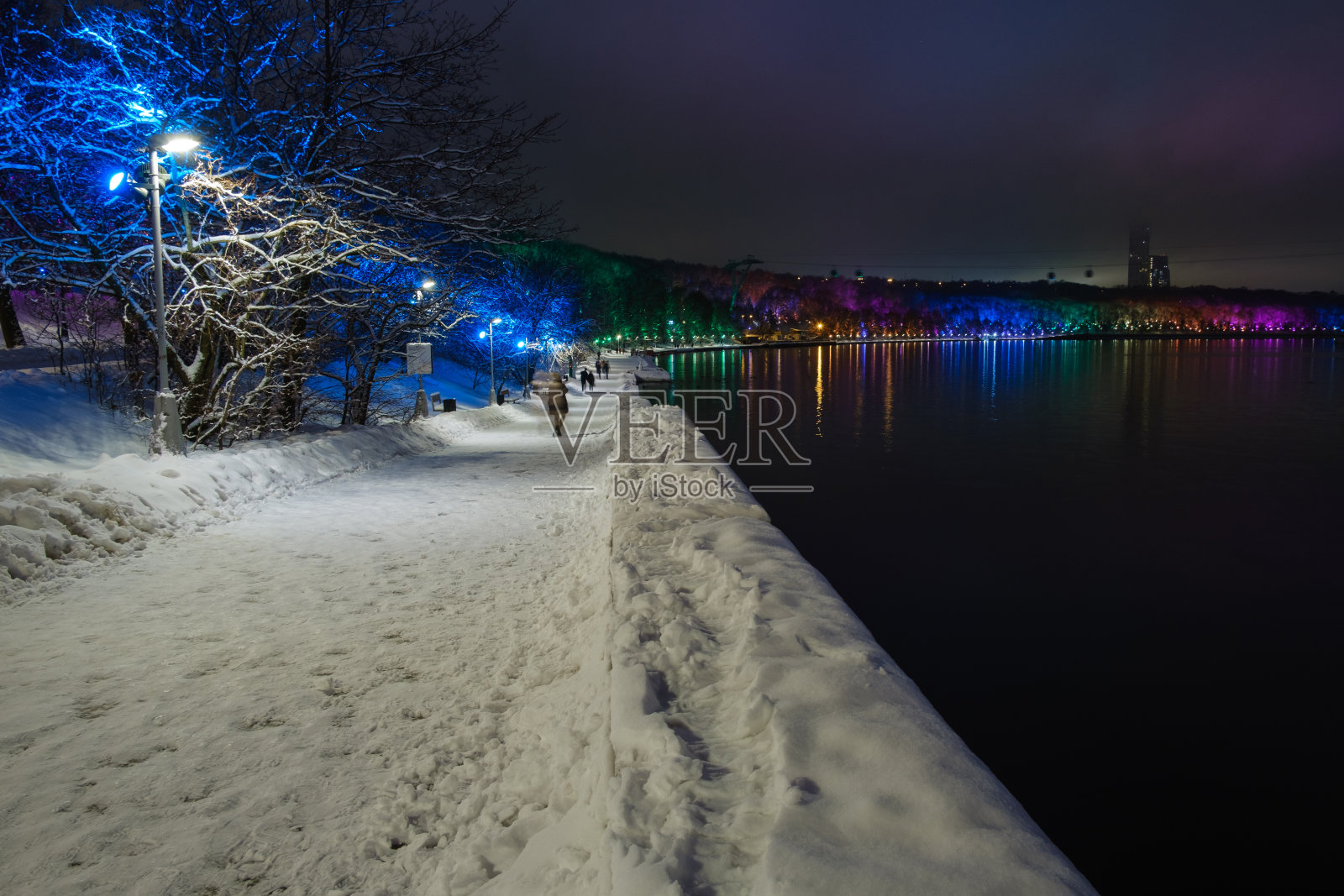 冬天的夜晚，公园里被白雪覆盖的河堤被彩色照明照亮照片摄影图片