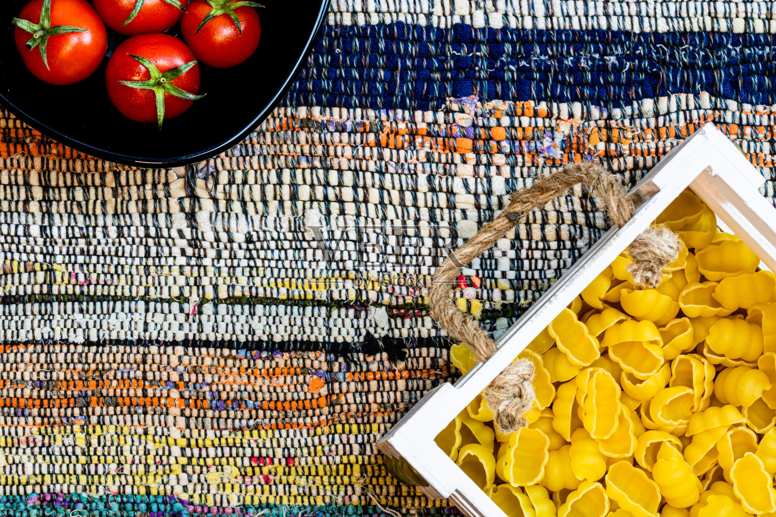 顶部细节的意大利面在木箱和新鲜成熟的樱桃番茄在一个黑色的碗上的乡村餐巾。配料和食物概念照片摄影图片