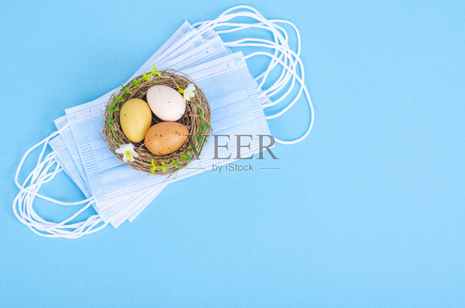 巢与明亮染色的鸡蛋与医疗口罩在蓝色背景，空格的文字。冠状病毒大流行期间的复活节假期照片摄影图片