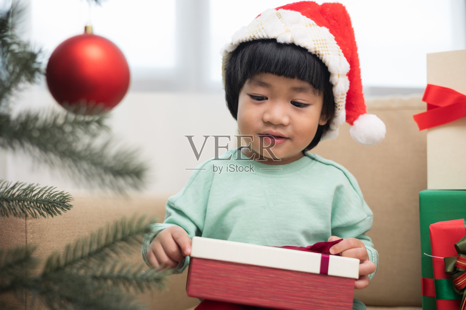 亚洲可爱的小男孩拿着礼物礼品盒，坐在客厅的沙发上，开心地装饰着圣诞树。可爱的男孩收到礼物。在圣诞快乐新年佳节。照片摄影图片