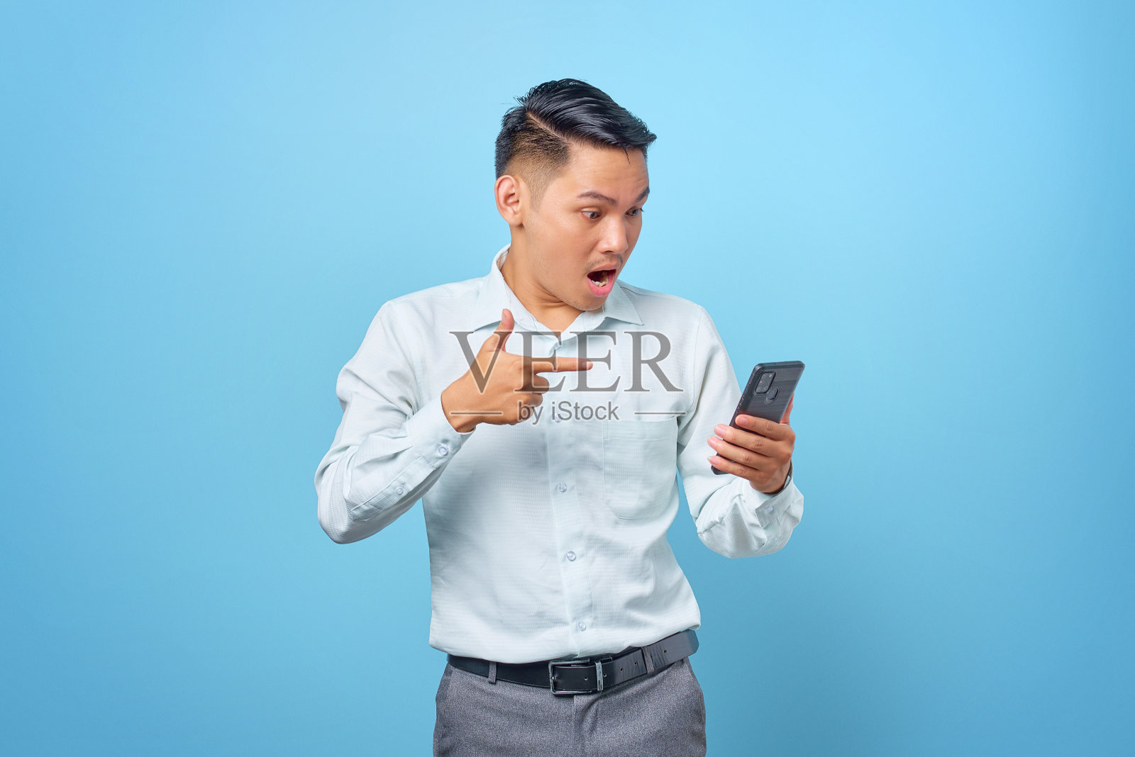 震惊的年轻英俊的商人指着蓝色背景下的智能手机照片摄影图片