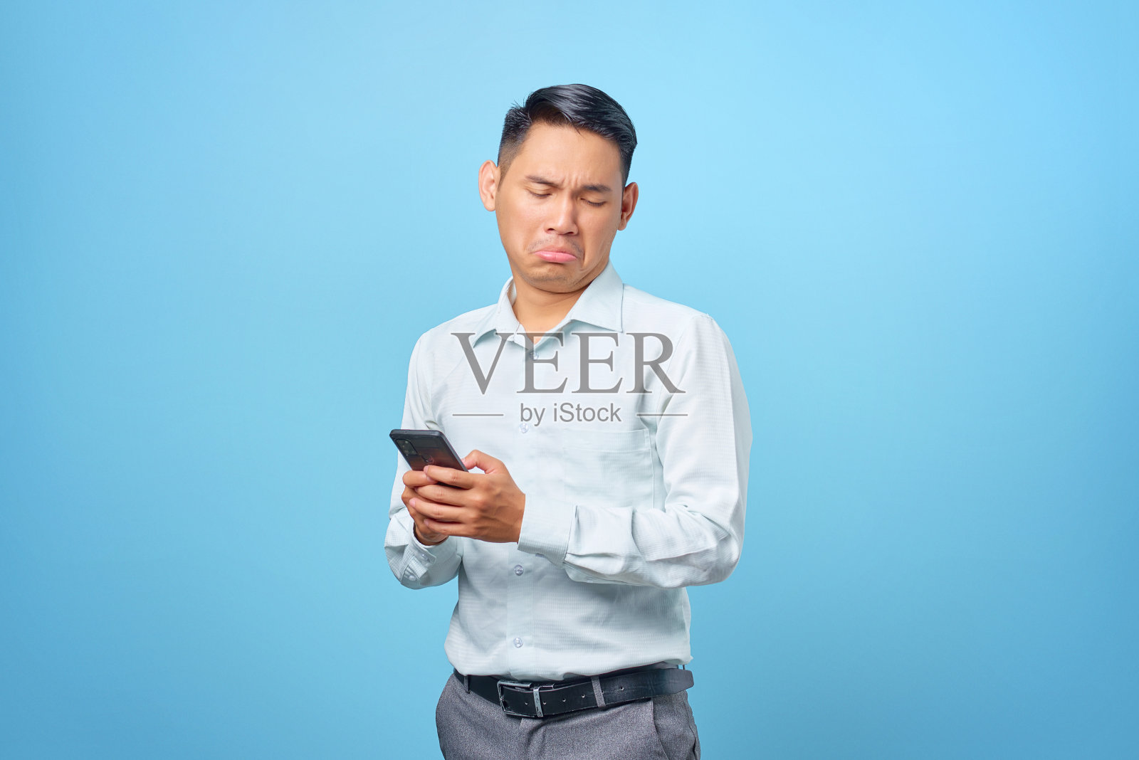 悲伤的年轻英俊的商人拿着并在蓝色背景下使用智能手机照片摄影图片