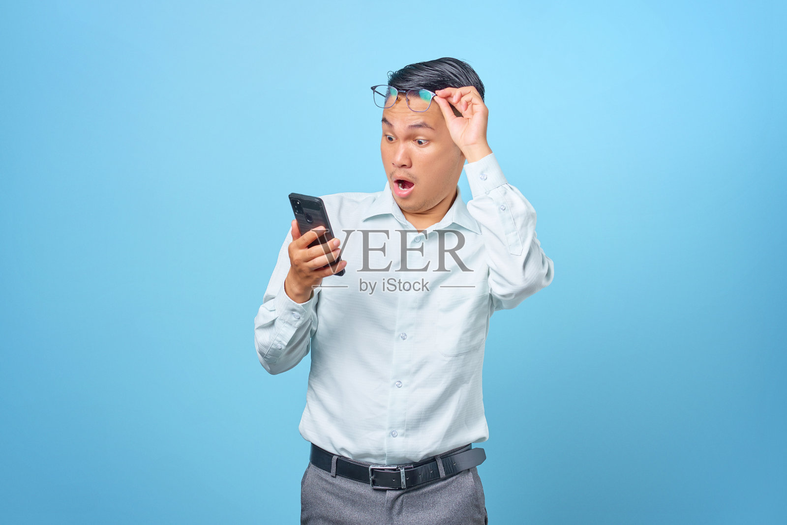 在蓝色背景下，年轻英俊的商人用智能手机摘掉眼镜，震惊了照片摄影图片
