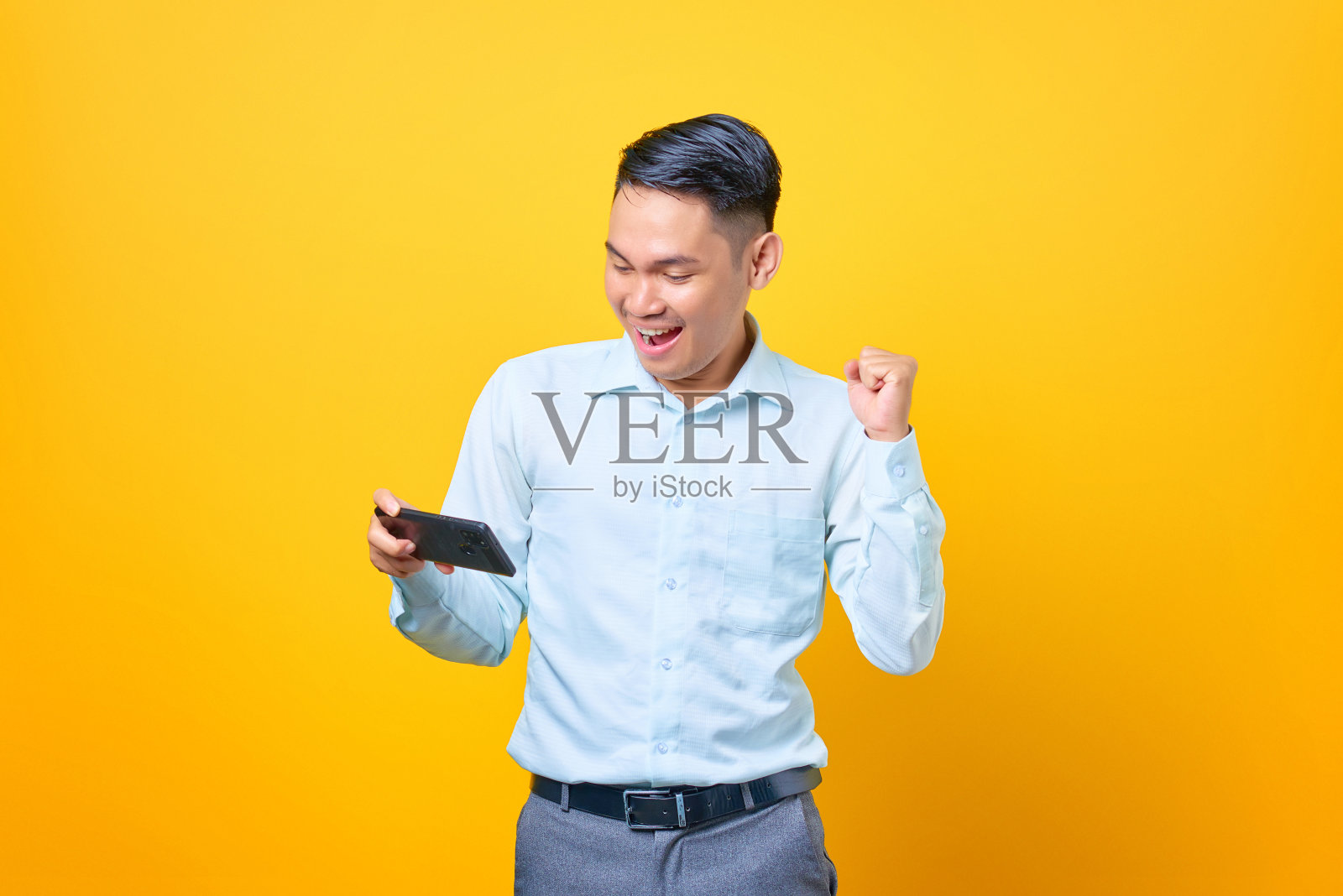 兴奋的年轻英俊的商人使用智能手机，在黄色背景上做胜利手势照片摄影图片