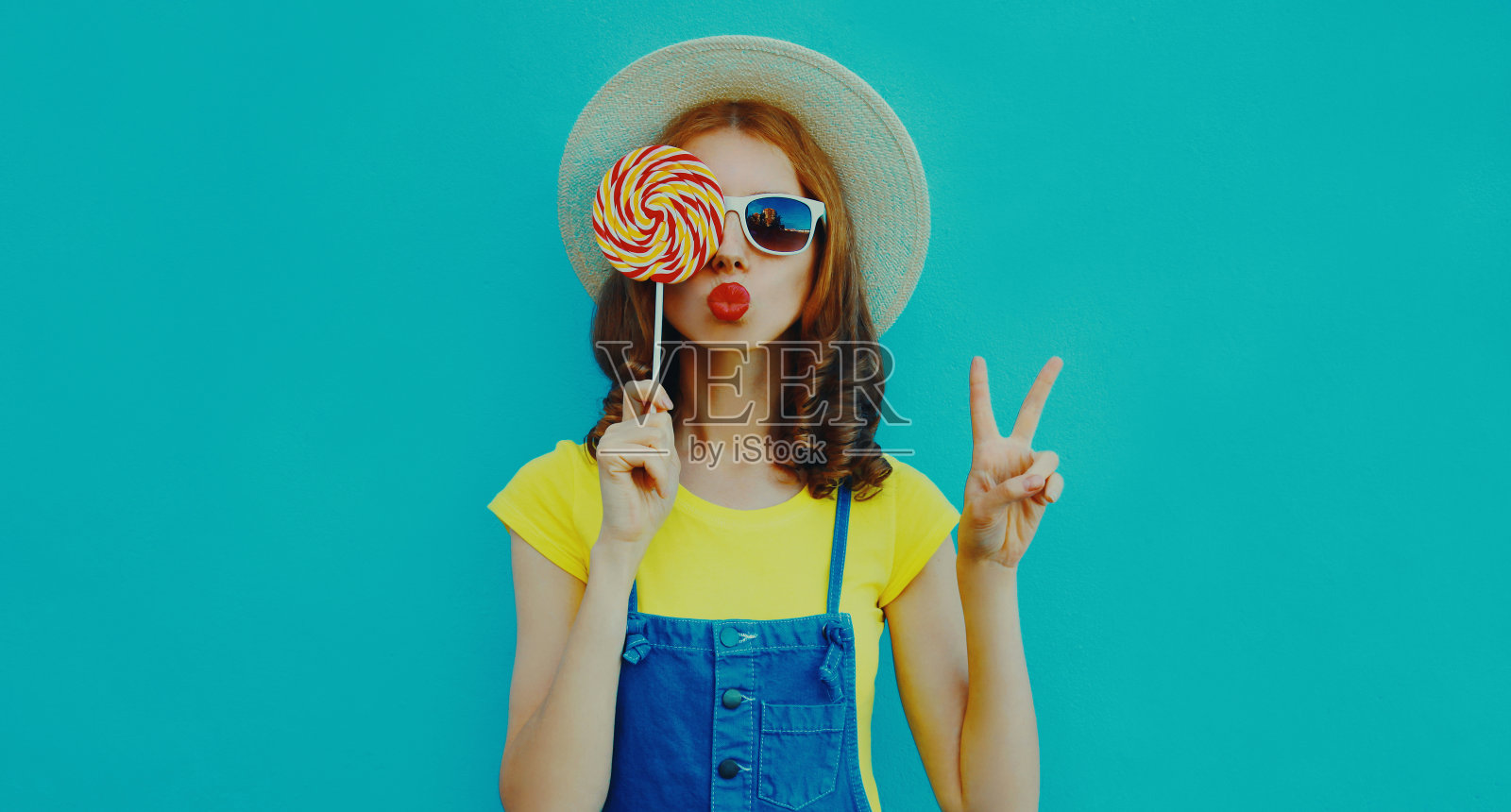 夏天的画像，美丽的年轻女子与棒棒糖吹她的嘴唇送甜蜜的空气吻穿着草帽在蓝色的背景照片摄影图片