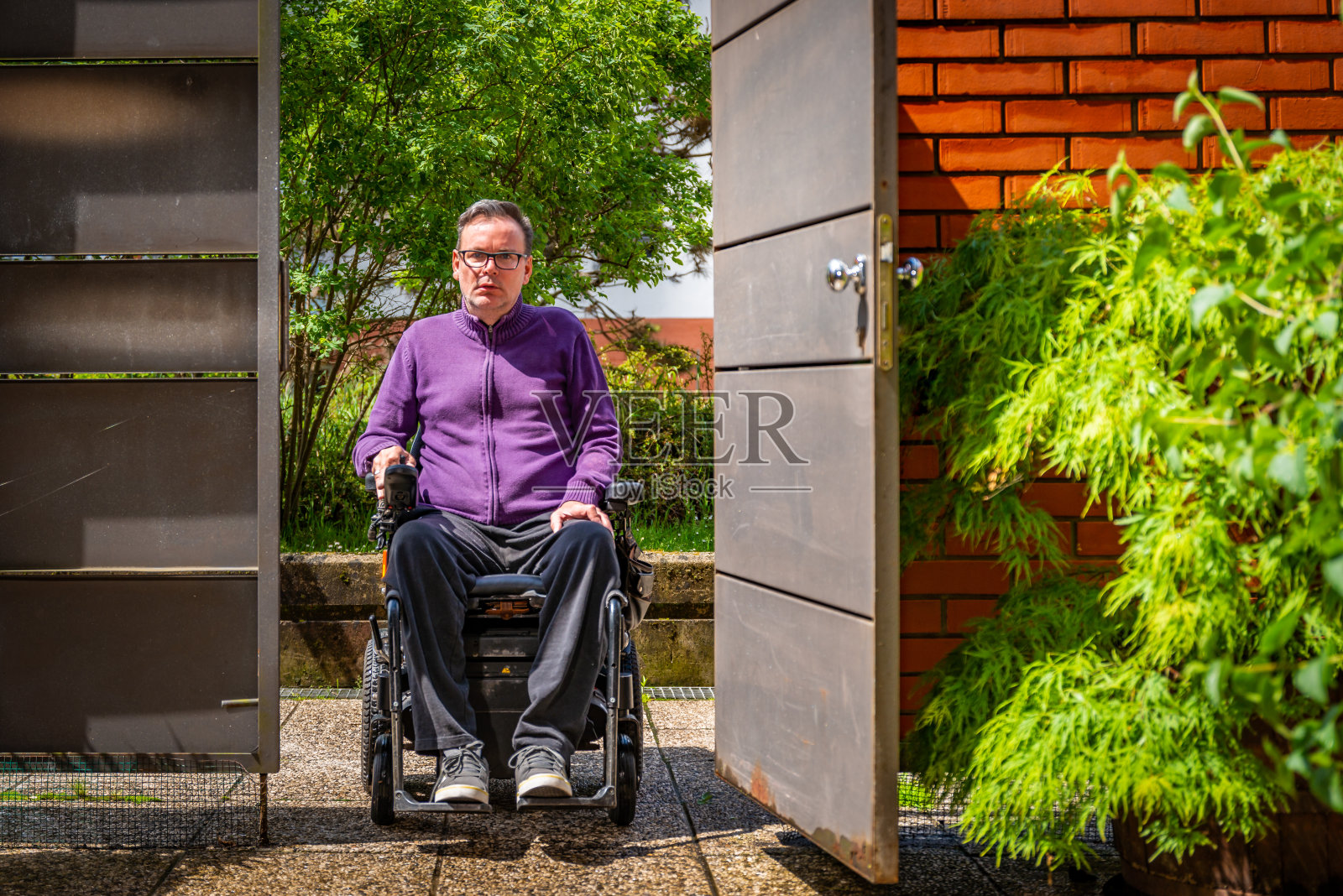 一名患有肌营养不良症的残疾男子坐在他庭院的轮椅上，身后是砖墙，周围是绿树，观察周围环境和绿化，进出照片摄影图片