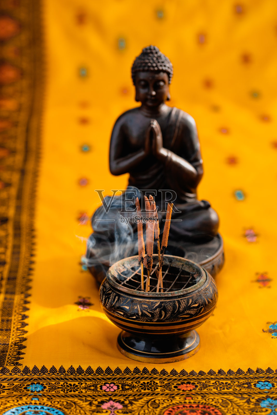 在阳光下，在一个没有聚焦的棕色佛像前，香碗里的熏香和带有刺绣的黄橙色印度教纱丽绳照片摄影图片