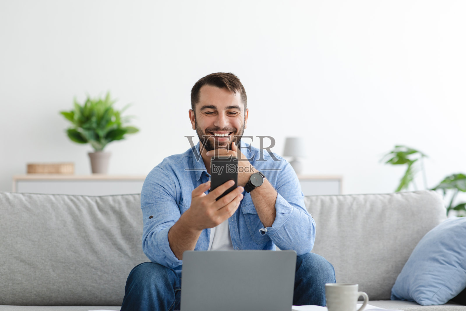 快乐的中年欧洲男性胡子在笔记本电脑远程工作，看手机在客厅内部照片摄影图片