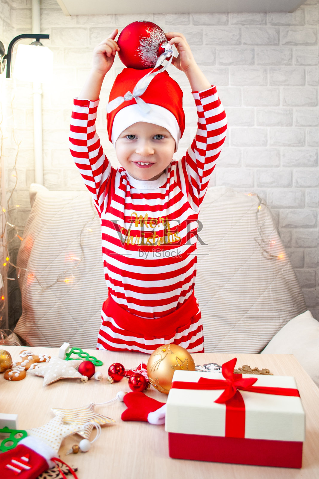 一个小孩子戴着圣诞老人的帽子，头上顶着一个红色的圣诞球，一边笑一边做鬼脸。男孩旁边是礼物照片摄影图片
