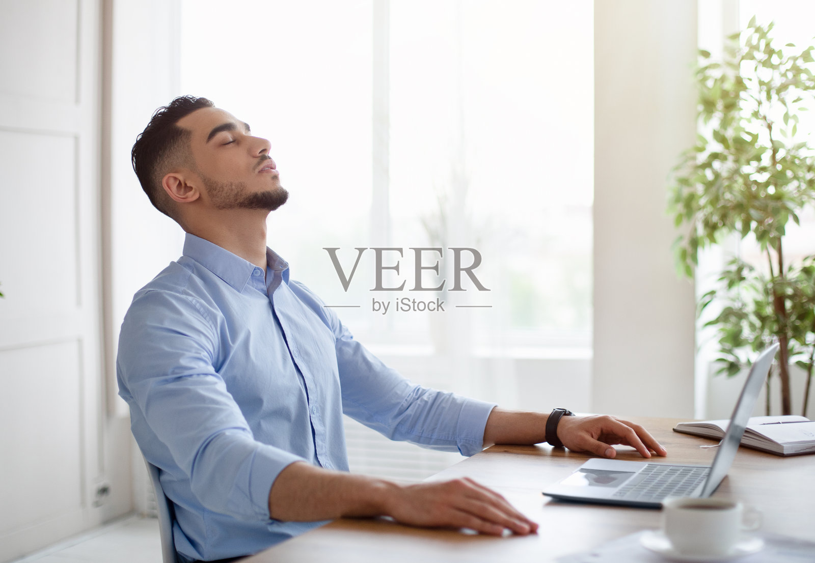 疲惫的年轻阿拉伯男子在现代办公室里闭着眼睛坐在笔记本电脑旁照片摄影图片