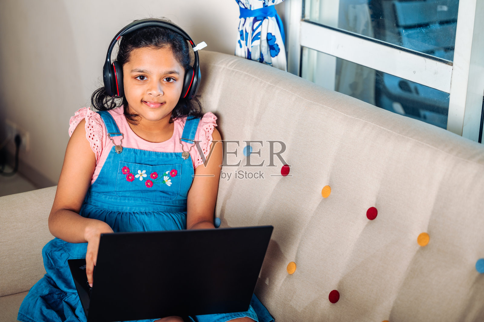 一个微笑的小女孩坐在家里的沙发上使用笔记本电脑。照片摄影图片
