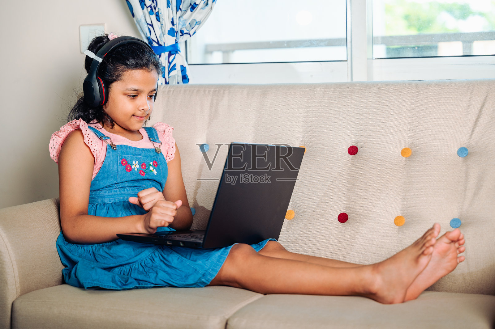 一个微笑的小女孩坐在家里的沙发上使用笔记本电脑。照片摄影图片